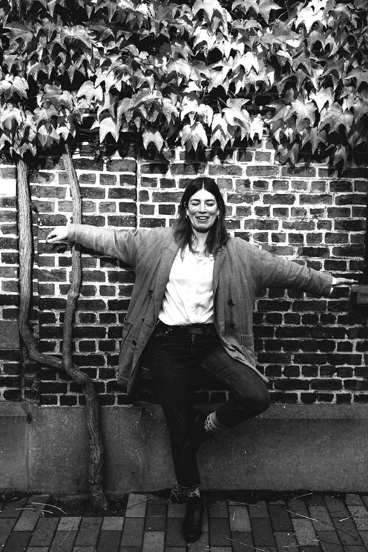 Christiane übt lächelnd die Yogahaltung Baum an einer Backsteinwand
